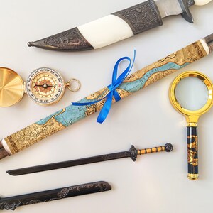 Puede incluir: Un surtido de art&iacute;culos de estilo antiguo, que incluyen una br&uacute;jula, un pergamino de mapa atado con una cinta azul, una lupa, un cuchillo decorativo y una espada peque&ntilde;a. Los art&iacute;culos est&aacute;n dispuestos sobre una superficie blanca.
