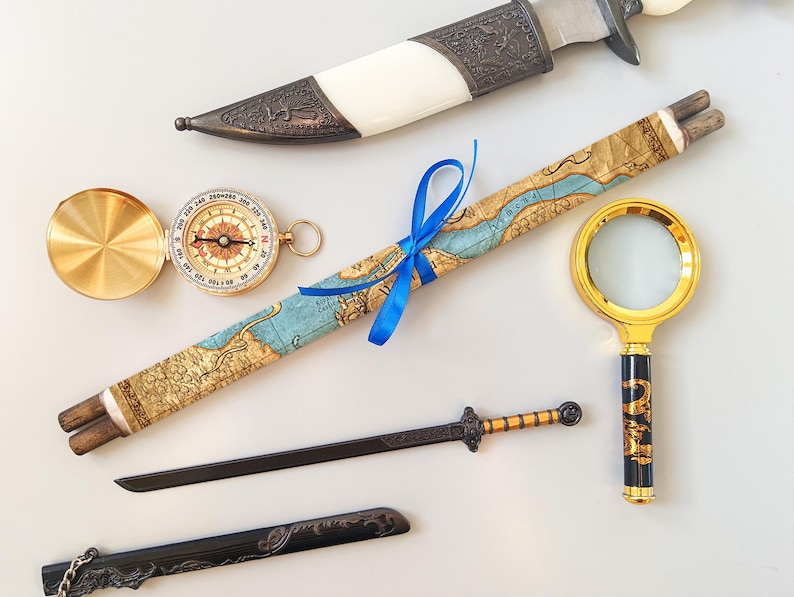 Puede incluir: Un surtido de art&iacute;culos de estilo antiguo, que incluyen una br&uacute;jula, un pergamino de mapa atado con una cinta azul, una lupa, un cuchillo decorativo y una espada peque&ntilde;a. Los art&iacute;culos est&aacute;n dispuestos sobre una superficie blanca.