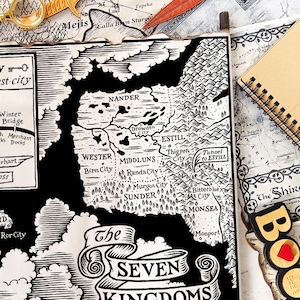 May include: Black and white fantasy map titled "The Seven Kingdoms" with detailed illustrations of cities and geographical features. Includes a magnifying glass, a decorative sword, and a wooden bookmark.