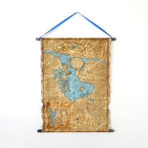 Puede incluir: Un mapa de Temeria, con una combinaci&oacute;n de colores azul y beige, cuelga de una barra de madera y una cinta azul. El mapa presenta una representaci&oacute;n detallada de una tierra con un gran lago y r&iacute;os. El texto "Temeria" est&aacute; en la parte superior del mapa.