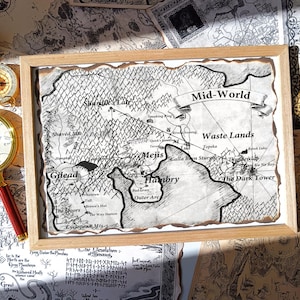 Op de afbeelding: Een ingelijste kaart van Mid-World met gedetailleerde geografische kenmerken en tekst. De kaart staat in een houten lijst, omringd door andere kaarten en een kompas. Twee vergrootglazen met rode handvatten en een gouden rand bevinden zich aan de zijkanten.