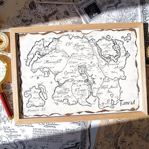 May include: A framed map of Tamriel, featuring detailed black ink illustrations of regions like Skyrim and Cyrodiil on aged paper. The map is set within a light wooden frame, with a compass and magnifying glass nearby.