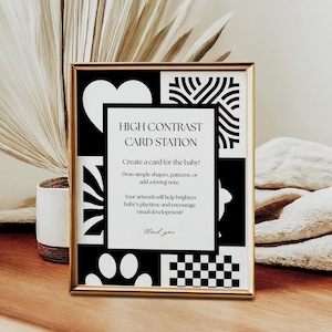 Puede incluir: Un letrero enmarcado en dorado con patrones geométricos en blanco y negro y el texto "HIGH CONTRAST CARD STATION". El letrero promueve la creación de tarjetas para bebés para mejorar el desarrollo visual. Una planta decorativa y tela doblada en el fondo.