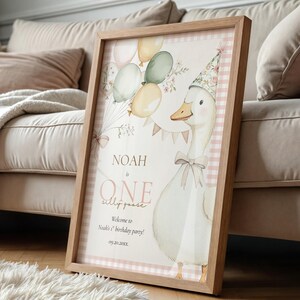May include: A framed print featuring a whimsical illustration of a goose wearing a party hat, balloons, and the text "Noah is One". The print has a light wood frame and a pink gingham border, perfect for a child's birthday party.