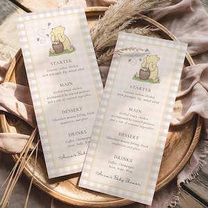 May include: Two baby shower menus with a Winnie the Pooh illustration. The menus have a beige and white checkered border and include the menu items: starter, main, dessert, and drinks. The text "Anna's Baby Shower" is at the bottom.