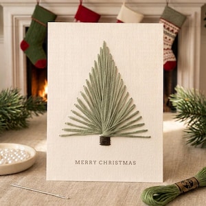 May include: A cream-colored greeting card featuring a hand-stitched Christmas tree made of sage green embroidery thread. The tree has a dark brown stitched trunk and the text MERRY CHRISTMAS is printed in a simple dark font at the bottom center.