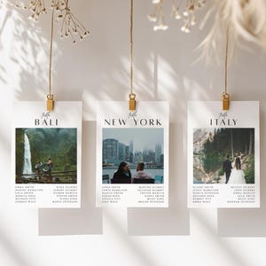 Puede incluir: Tres tarjetas de mesa de boda suspendidas por clips dorados. Cada tarjeta presenta un destino de viaje diferente: Bali, Nueva York e Italia. Cada tarjeta tiene una foto y una lista de nombres. Las tarjetas cuelgan sobre un fondo neutro.