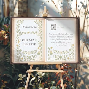 Pode incluir: Um livro aberto em um cavalete de madeira, com texto e ilustrações. A página esquerda diz "Welcome to OUR NEXT CHAPTER" com desenhos florais e mamadeiras. A página direita começa "Once upon a time..." e conta uma história sobre um bebê.