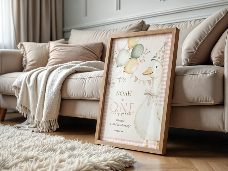 May include: A framed print featuring a whimsical illustration of a goose wearing a party hat, balloons, and the text "Noah is One". The print has a light wood frame and a pink gingham border, perfect for a child's birthday party.