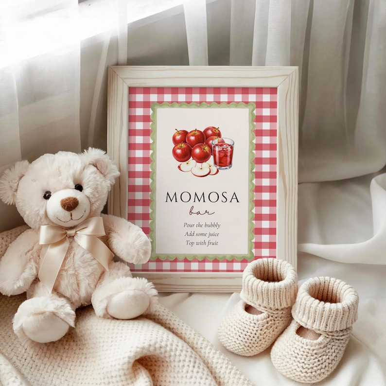 Op de afbeelding: Een ingelijst bord met een rood-witte geruite achtergrond en de woorden "MOMOSA bar". Het bord toont een illustratie van appels en een drankje. Een witte teddybeer, een deken en babyslofjes staan ook op de afbeelding.