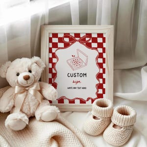 May include: A framed sign with a red and white chequered background and a red ribbon design. The sign features the words "CUSTOM sign" and "WRITE ANY TEXT HERE!". A teddy bear, a blanket, and two pairs of baby booties are in the image.