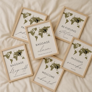 May include: A collection of framed signs with a travel theme. Each sign features a world map in olive green, a black airplane graphic, and text such as "Baggage Claim" and "Welcome Aboard" in elegant script. The frames are light wood.