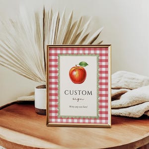 Op de afbeelding: Een ingelijst bord met een rood-witte ruitjesrand. Het bord toont een aquarel illustratie van een rode appel met een groen blad. De tekst "CUSTOM sign" staat onder de appel, met ruimte om aangepaste tekst te schrijven.