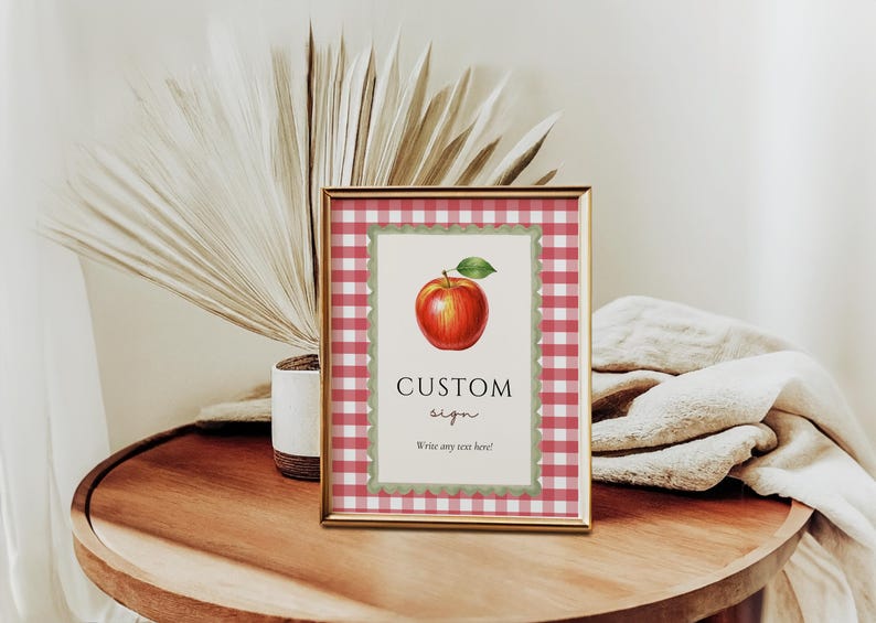 Op de afbeelding: Een ingelijst bord met een rood-witte ruitjesrand. Het bord toont een aquarel illustratie van een rode appel met een groen blad. De tekst "CUSTOM sign" staat onder de appel, met ruimte om aangepaste tekst te schrijven.