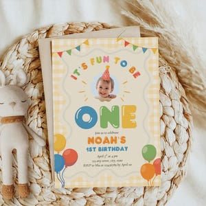 May include: A birthday invitation with the text "It's Fun to Be One" and "Noah's 1st Birthday". The invitation features a photo of a baby, balloons, and a decorative border. A stuffed bunny is next to the invitation.
