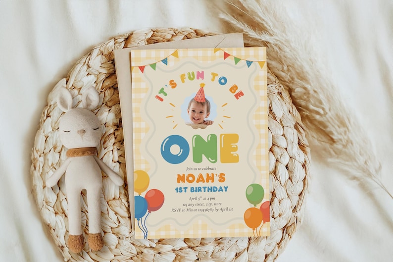May include: A birthday invitation with the text "It's Fun to Be One" and "Noah's 1st Birthday". The invitation features a photo of a baby, balloons, and a decorative border. A stuffed bunny is next to the invitation.