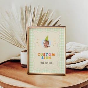May include: A framed custom sign with a baby wearing a party hat. The sign has a light green gingham border and the words "CUSTOM SIGN" in colorful letters. The text "YOUR TEXT HERE" is below.