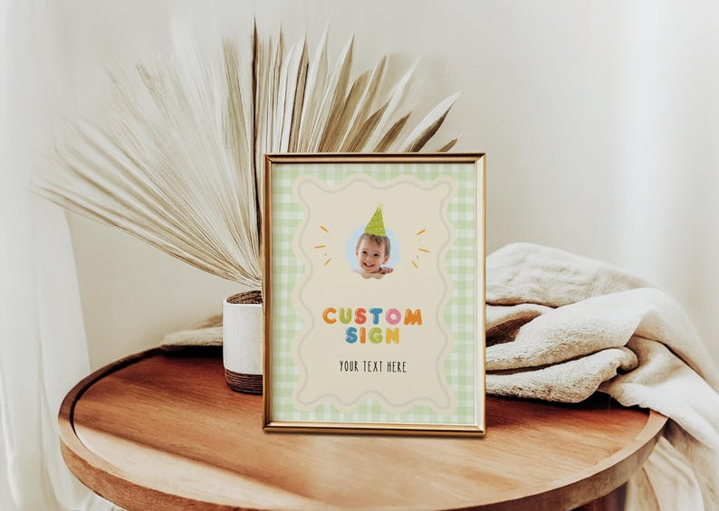 May include: A framed custom sign with a baby wearing a party hat. The sign has a light green gingham border and the words "CUSTOM SIGN" in colorful letters. The text "YOUR TEXT HERE" is below.