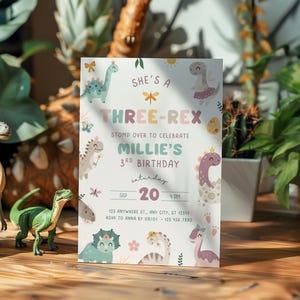 Pode incluir: Um convite de anivers&aacute;rio com tema de dinossauro. O convite diz "She's a Three-Rex" e celebra o 3&ordm; anivers&aacute;rio de Millie. O design apresenta dinossauros de desenho animado em cores pastel. Duas figuras de dinossauros est&atilde;o em primeiro plano.