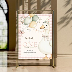May include: A party sign featuring a cartoon goose wearing a floral party hat and bow tie, holding balloons. The sign reads "Noah is One silly goose" with a welcome message for Noah's first birthday party. The background has a pink gingham pattern.
