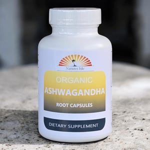 Op de afbeelding: Witte fles Natures Isle Organic Ashwagandha Root Capsules. Het etiket heeft een geel en wit ontwerp met de tekst "Organic Ashwagandha Root Capsules" en "Voedingssupplement". De bovenkant van het etiket heeft een logo van een palmboom en de zon.