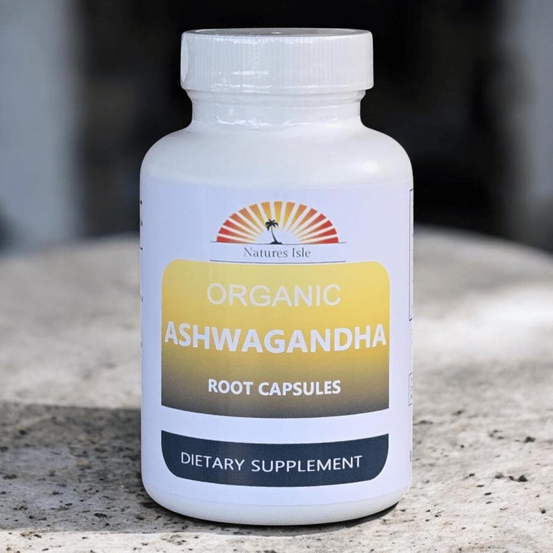 Op de afbeelding: Witte fles Natures Isle Organic Ashwagandha Root Capsules. Het etiket heeft een geel en wit ontwerp met de tekst "Organic Ashwagandha Root Capsules" en "Voedingssupplement". De bovenkant van het etiket heeft een logo van een palmboom en de zon.