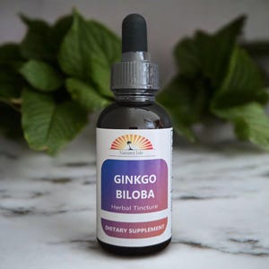 Puede incluir: Una botella de vidrio oscuro de tintura herbal de Ginkgo Biloba con un cuentagotas negro. La etiqueta presenta un logotipo de puesta de sol, las palabras "Ginkgo Biloba" y "Suplemento dietético."