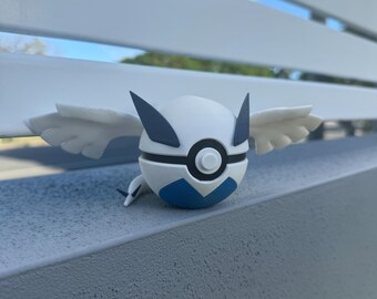 Może przedstawiać: Biała i niebieska figurka Pokemon z skrzydłami. Figurka jest kulą z czarnym kółkiem i białym przyciskiem z przodu. Figurka stoi na szarej powierzchni.