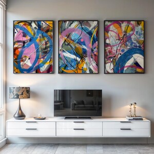 Könnte beinhalten: Drei gerahmte abstrakte Kunstwerke mit schwarzen Rahmen. Jedes Kunstwerk zeigt eine lebendige Mischung aus Farben, darunter Rosa, Blau, Gelb und Orange, mit dynamischen Pinselstrichen und Linien. Die Kunst wird über einem weißen Schrank mit einem Fernseher ausgestellt.