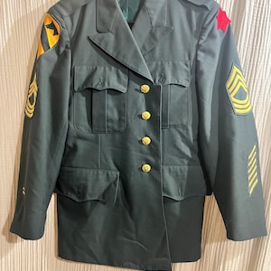 Op de afbeelding: Een donkergroen militair uniformjack met gouden knopen en verschillende patches op de mouwen. De patches omvatten een rode ster, een gele chevron en een geel embleem.