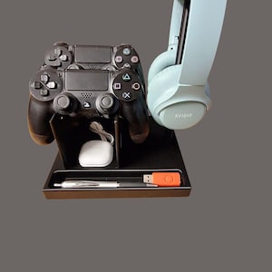 May include: A black charging station with two black video game controllers and a light blue headset with the word "KV1010" printed on the side. The station has a small white charging puck and a black pen.