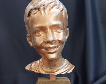 May include: A bronze-colored sculpture of a smiling boy's head and shoulders. The bust is mounted on a wooden base. The sculpture has a metallic finish with visible brushstrokes. The base is made of rough-hewn wood.