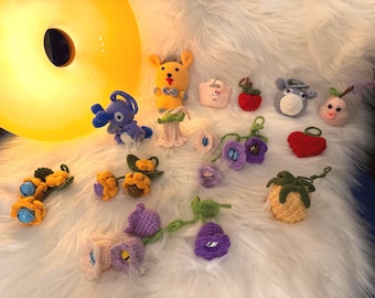 Può includere: Una collezione di figure amigurumi e disegni floreali colorati all'uncinetto. L'assortimento comprende un personaggio giallo simile a Pikachu, un elefante blu, una mela rossa e vari fiori. Gli oggetti sono disposti su una superficie bianca e soffice.