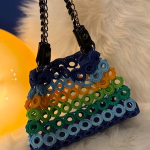 May include: A small, colorful crochet bag with a chain strap. The bag features a rainbow of colors, including blue, green, yellow, and orange, arranged in a circular pattern. The strap is a black chain with black leather accents.