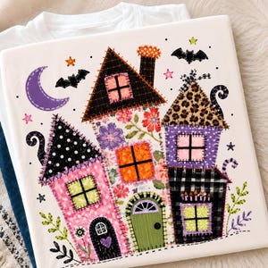 May include: A white t-shirt featuring a colorful graphic of a patchwork-style house with various patterns including polka dots, leopard print, plaid, and floral designs. The illustration includes a purple crescent moon, black bats, and stars.