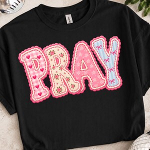 Könnte beinhalten: Ein schwarzes T-Shirt mit dem Wort PRAY in großen, dekorativen Buchstaben. Jeder Buchstabe hat ein eigenes Muster: rosa Vichy-Karo mit Herzen, cremefarbener Blumendruck, rosa Schneeflocken-Design und hellblaue Streifen mit kleinen Schleifen.
