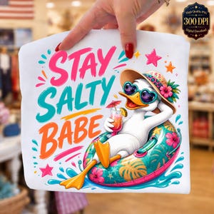 Könnte beinhalten: Weißes T-Shirt mit einem farbenfrohen Aufdruck einer Ente mit Sonnenbrille und Sonnenhut, die sich auf einem aufblasbaren Ring entspannt. Der Text "STAY SALTY BABE" ist in Pink, Türkis und Orange dargestellt.