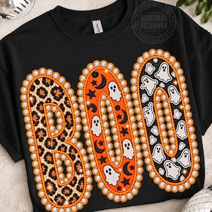 May include: A black crew neck t-shirt featuring the word BOO in large letters. The B has a leopard print, the O has an orange background with black stars, moons, and white ghosts, and the final O has a black background with white ghosts.