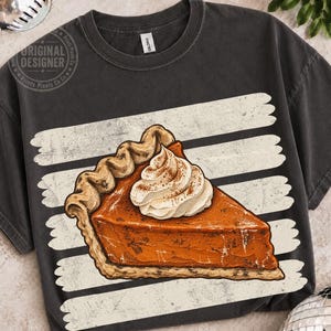 Op de afbeelding: Een antracietgrijs T-shirt met ronde hals en een afbeelding van een stuk pompoentaart met een toef slagroom. De taart is gecentreerd over een reeks horizontale, verweerde witte penseelstreeplijnen op de voorkant van het shirt.