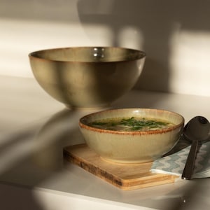 Può includere: Due ciotole in ceramica verde oliva, una riempita di zuppa e guarnita con erbe fresche. Le ciotole hanno un aspetto rustico e strutturato con un bordo marrone. Sono visibili anche un tagliere di legno e un cucchiaio.