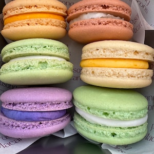 May include: A box of colorful macarons in various shades, including orange, pink, green, yellow, purple, and white. The macarons are arranged in rows, with visible fillings between the cookie shells. The box has the text "Pusong Macarons".