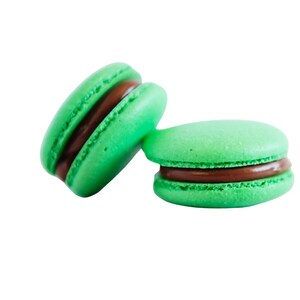 K&ouml;nnte beinhalten: Zwei gr&uuml;ne Macarons mit dunkler Schokoladenf&uuml;llung vor wei&szlig;em Hintergrund. Die Macarons sind rund, mit leicht strukturierter Oberfl&auml;che. Eines der Macarons lehnt am anderen und zeigt die F&uuml;llung. Ein s&uuml;&szlig;er Genuss.