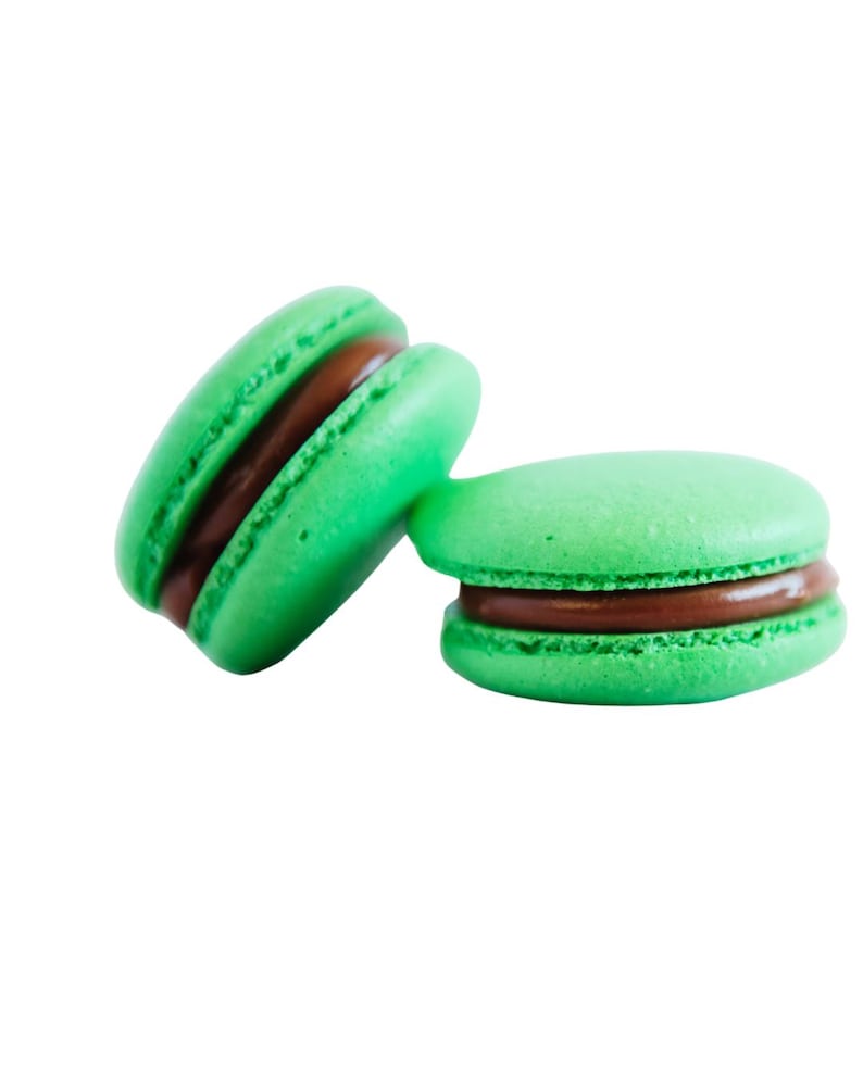 K&ouml;nnte beinhalten: Zwei gr&uuml;ne Macarons mit dunkler Schokoladenf&uuml;llung vor wei&szlig;em Hintergrund. Die Macarons sind rund, mit leicht strukturierter Oberfl&auml;che. Eines der Macarons lehnt am anderen und zeigt die F&uuml;llung. Ein s&uuml;&szlig;er Genuss.