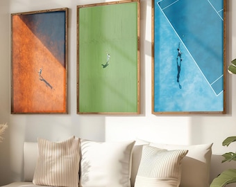 May include: Three framed tennis court art prints. One print features an orange court, another a green court, and the third a blue court. Each print has a wooden frame and depicts a tennis player. The prints are displayed above a white sofa.