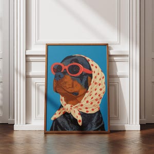 Puede incluir: Una impresi&oacute;n art&iacute;stica enmarcada que presenta a un perro Rottweiler con gafas de sol rojas y un pa&ntilde;uelo crema con lunares rojos. El perro est&aacute; sobre un fondo azul brillante. La obra de arte est&aacute; en un marco de madera.