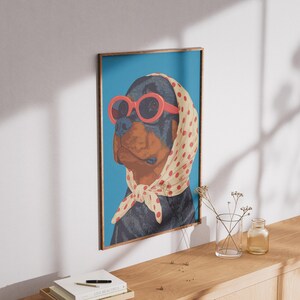 Puede incluir: Una impresi&oacute;n art&iacute;stica enmarcada con un perro Rottweiler que lleva gafas de sol rojas y un pa&ntilde;uelo crema con lunares rojos. El perro est&aacute; sobre un fondo azul. El marco es marr&oacute;n.