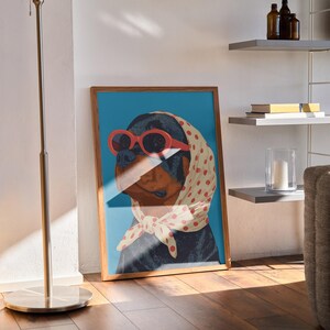 Puede incluir: Impresi&oacute;n art&iacute;stica enmarcada que presenta un perro Rottweiler con gafas de sol rojas y un pa&ntilde;uelo crema con lunares rojos. El perro est&aacute; sobre un fondo azul. La obra de arte est&aacute; en un marco de madera.