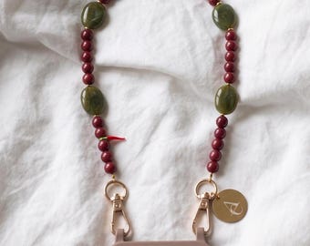 Può includere: Un cordino per telefono con perline sferiche color marrone e perline ovali verde oliva, arricchito da un piccolo ciondolo a forma di peperoncino rosso. Si fissa a una cover color malva tramite moschettoni dorati e una medaglietta dorata con logo bianco.