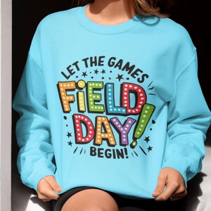 May include: A light blue sweatshirt with the text "Let the games begin!" and "Field Day!" in colorful, outlined letters with light bulbs around them.