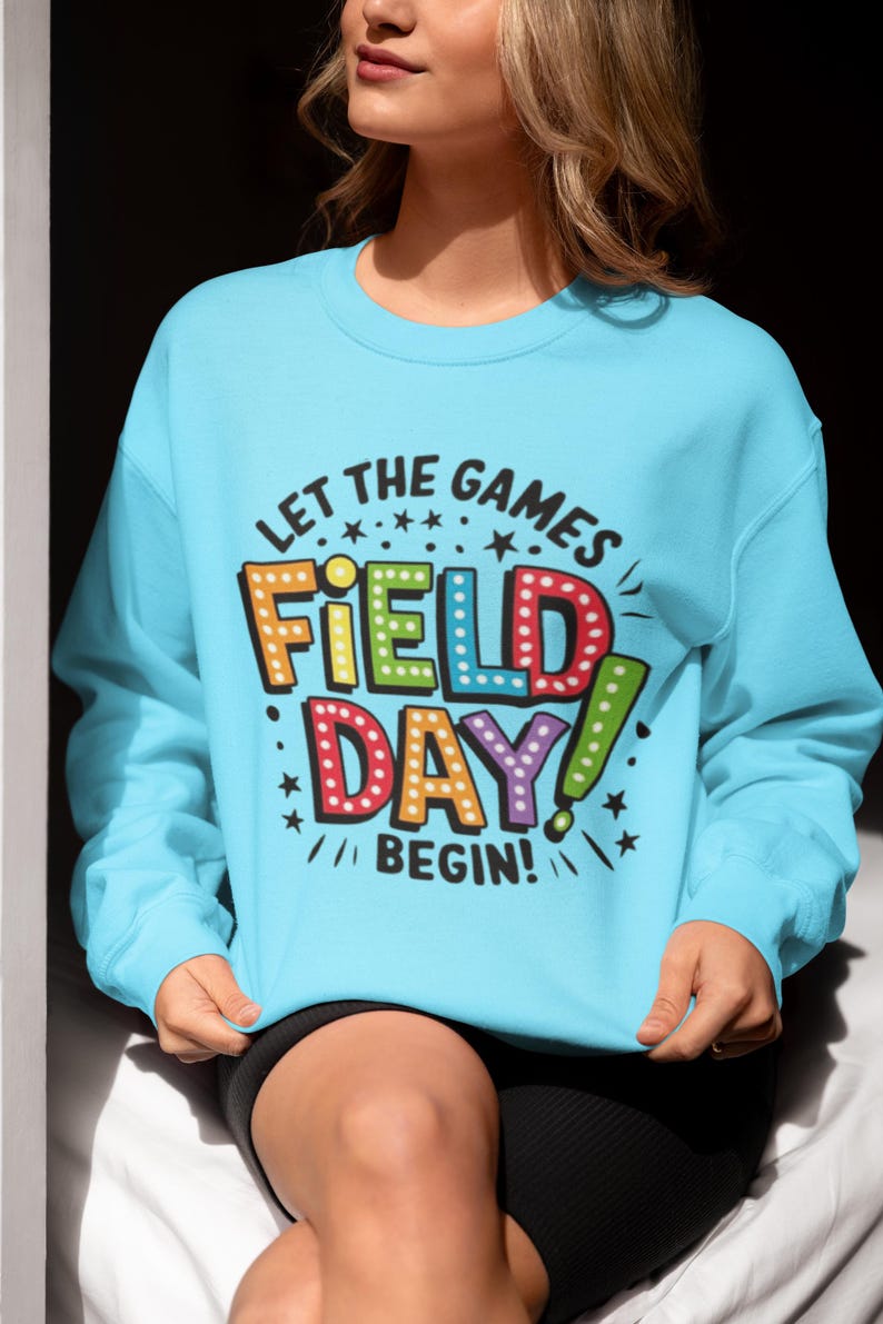 May include: A light blue sweatshirt with the text "Let the games begin!" and "Field Day!" in colorful, outlined letters with light bulbs around them.